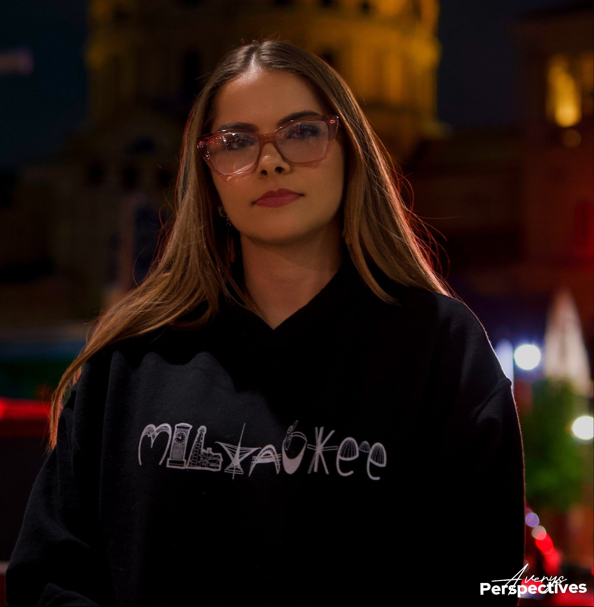 Mariah Grace wearing a black sweatshirt with 'Milwaukee' landmarks text in front of a blurred building at night.